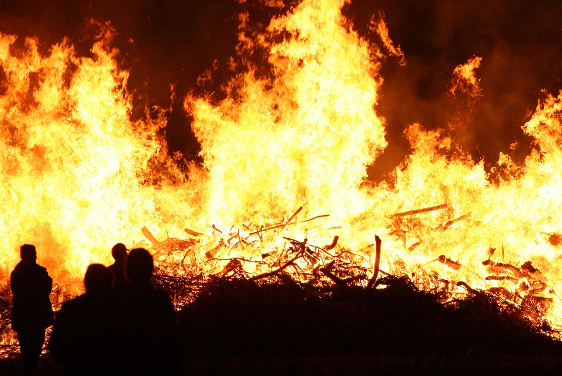 En brinnande Valborgsmässo eld med mörka siluetter av människor i förgrunden.